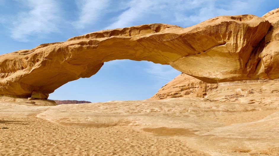 Brown rock shaped like a bridge over brown ground in front of a blue and slightly cloudy sky background