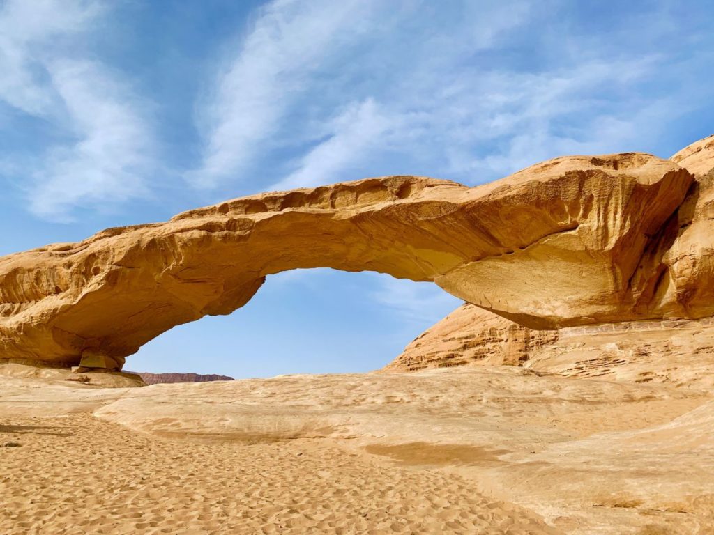 Brown rock shaped like a bridge over brown ground in front of a blue and slightly cloudy sky background