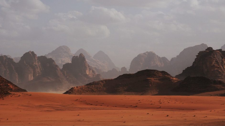 Wide view of red ground and brown rocky hills with a gray sky