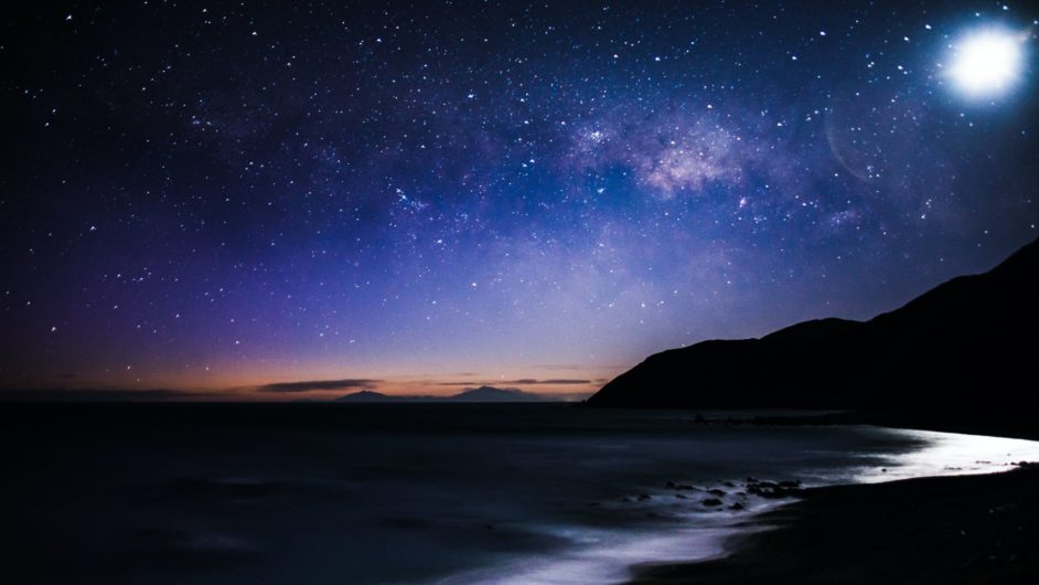 Landscape with water along a shoreline, hills in the background, a starry sky, and orange rays of light on the horizon